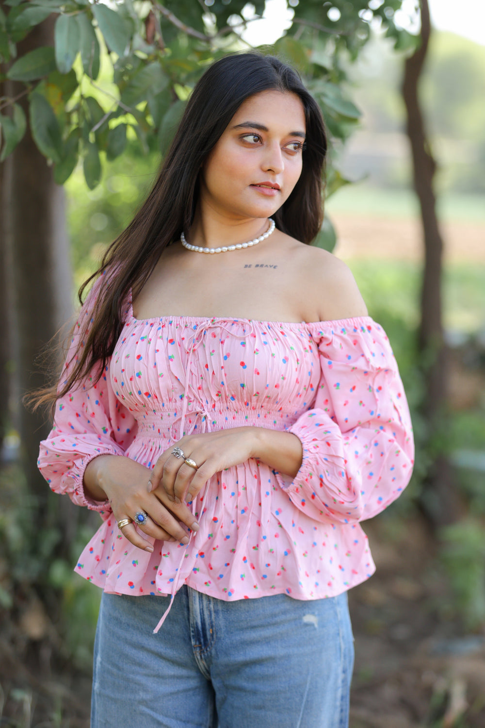 A woman wearing a pink sleeveless top with an open square neckline and a smocked empire waistband, standing in a garden.