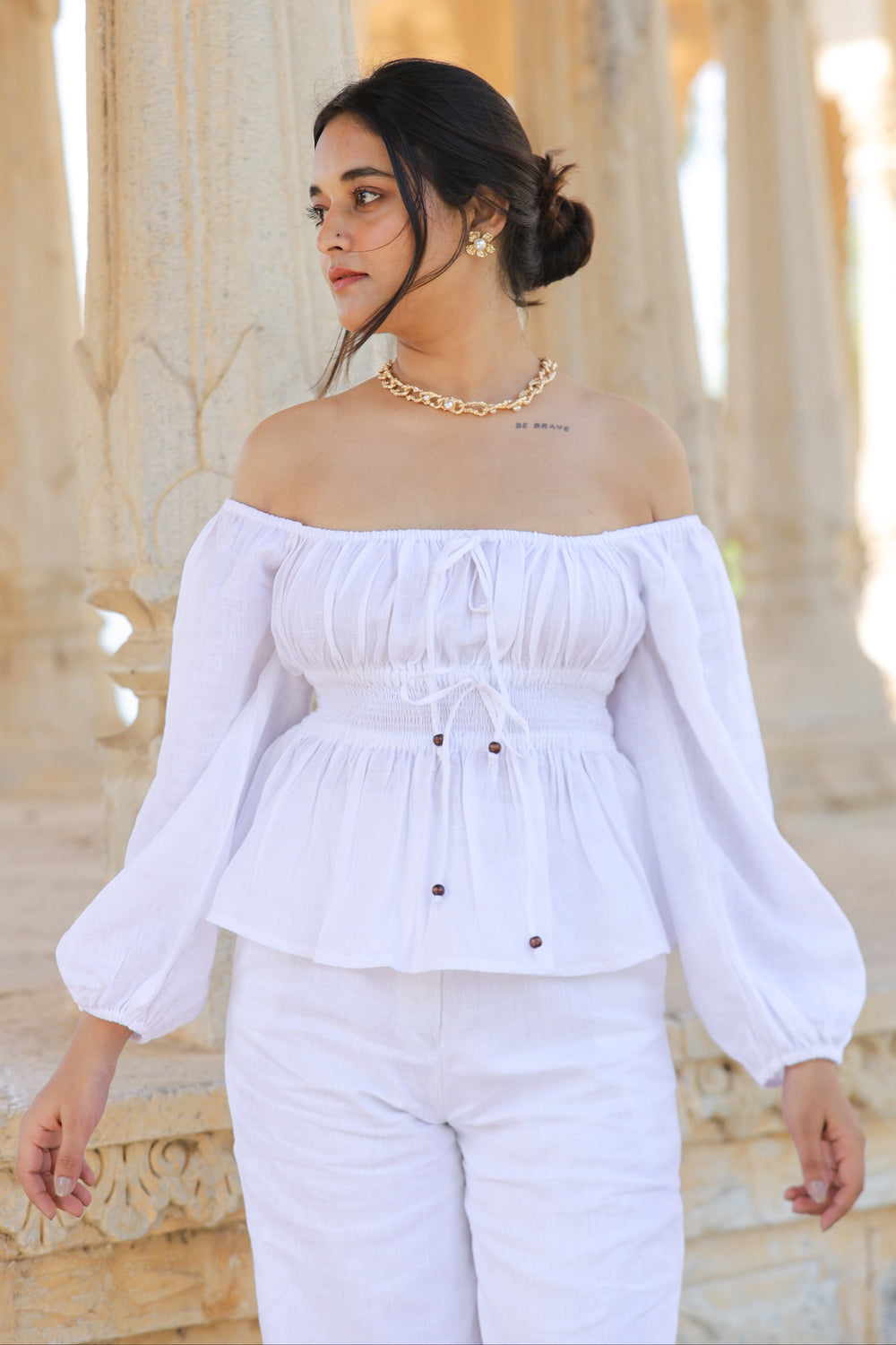 A woman wearing a white sleeveless top with an open square neckline and a smocked empire waistband, standing in a columned hallway.