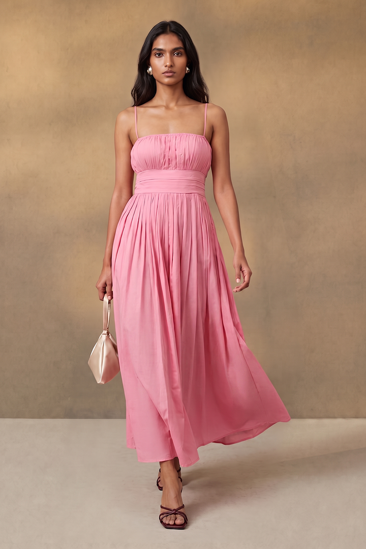 A woman modeling a pink spaghetti-strap maxi dress with a ruched bodice and flowing skirt, standing against a textured beige studio backdrop and holding a small cream handbag.