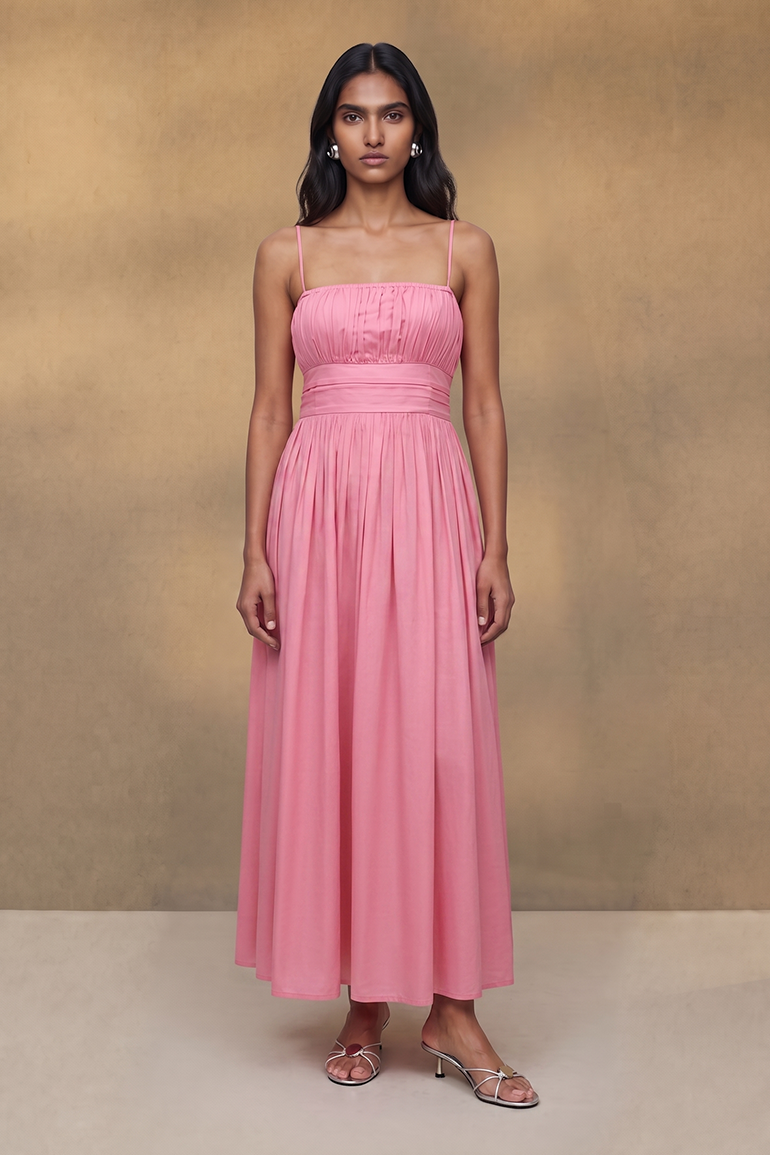 A woman stands facing forward wearing a long pink spaghetti-strap dress with a gathered bodice and full flowing skirt, photographed against a warm beige studio backdrop.