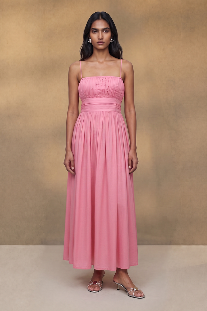 A woman stands facing forward wearing a long pink spaghetti-strap dress with a gathered bodice and full flowing skirt, photographed against a warm beige studio backdrop.