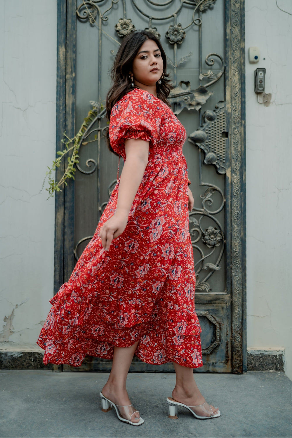 A woman wearing a red and white floral midi dress with puff sleeves and clear high-heeled sandals, standing in front of a weathered dark metal door.