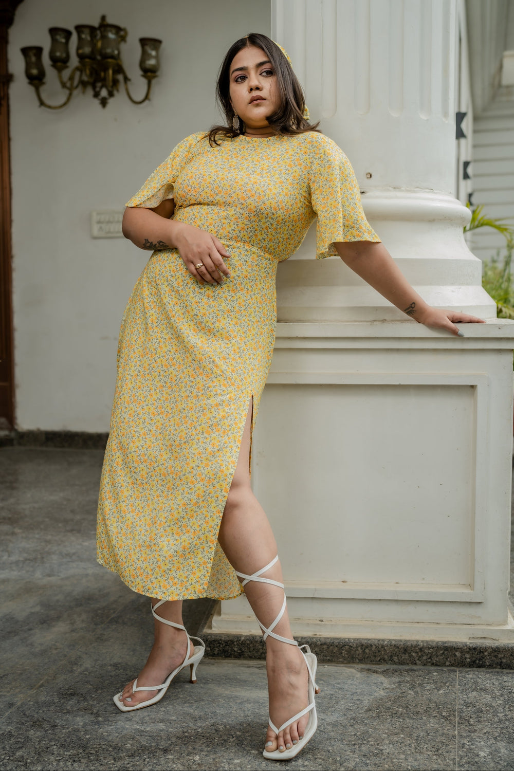 A woman models a yellow and white floral print midi dress with short flutter sleeves and a side slit, posing outdoors next to a white architectural column.