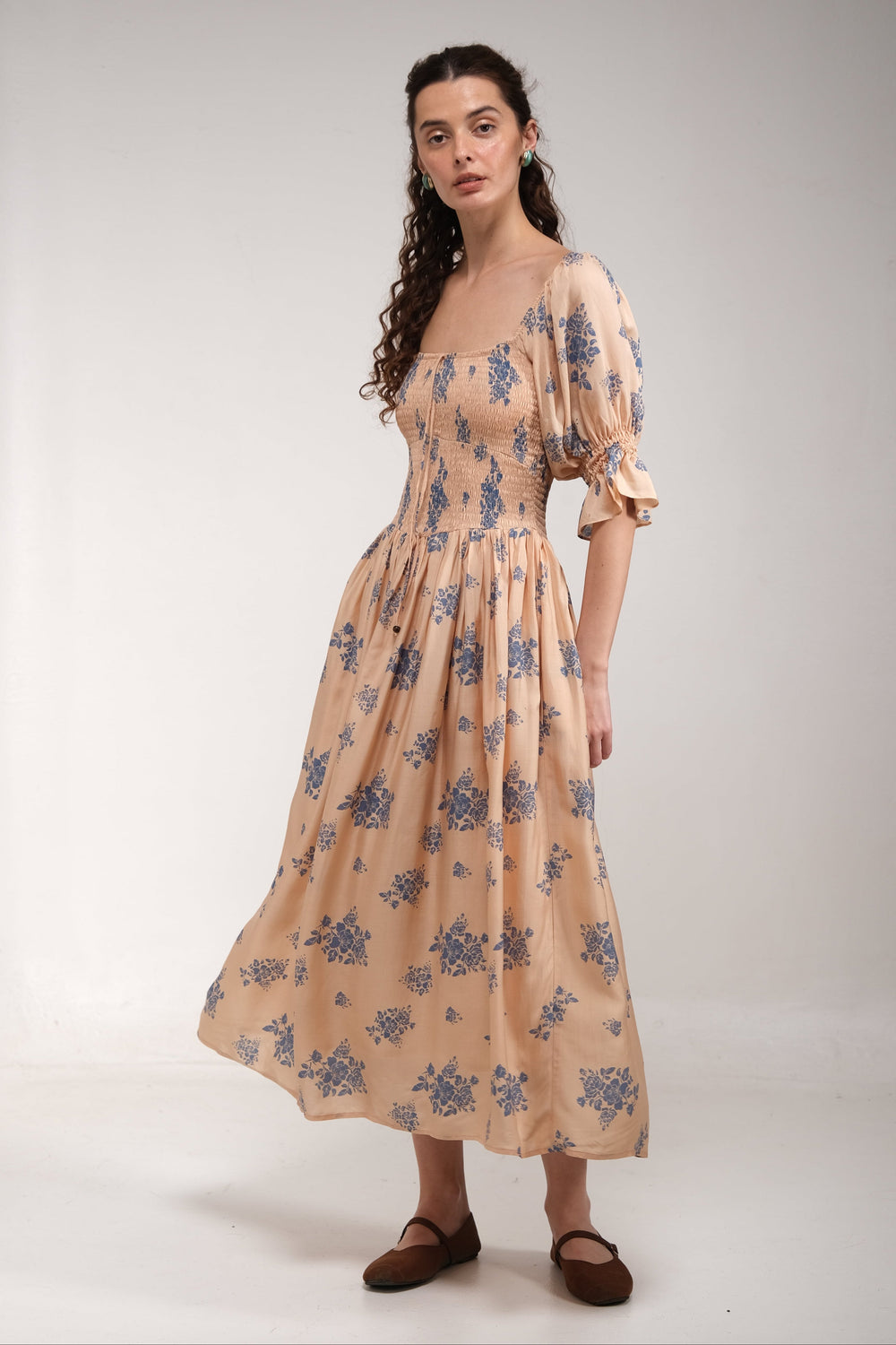 A woman modeling a midi-length beige dress with a blue floral pattern, featuring puff sleeves and a square neckline, set against a plain light gray studio background.