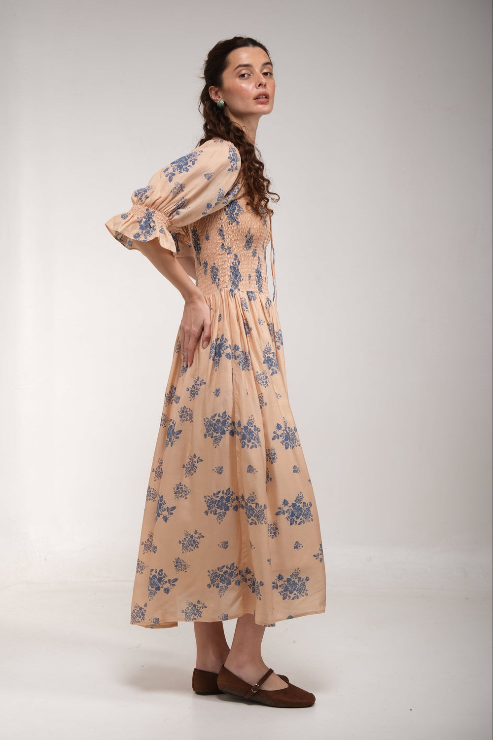 A woman modeling a long-sleeved midi dress with a blue floral pattern on a light beige background, photographed against a plain white studio backdrop.