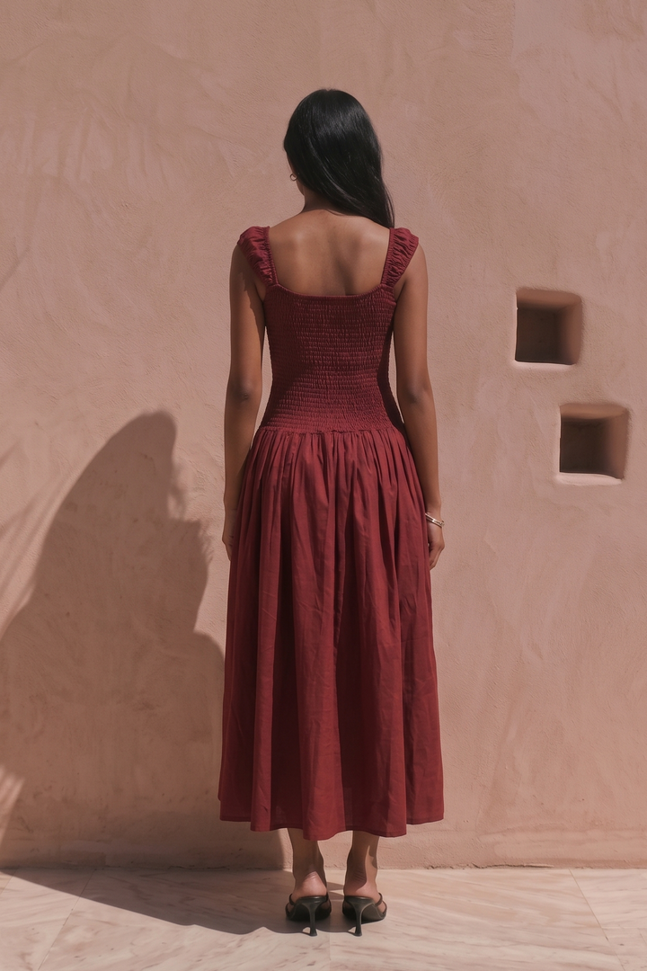 A woman stands with her back to the camera wearing a sleeveless rust-red midi dress with a smocked bodice and gathered skirt, set against a textured beige stucco wall with soft palm shadows.
