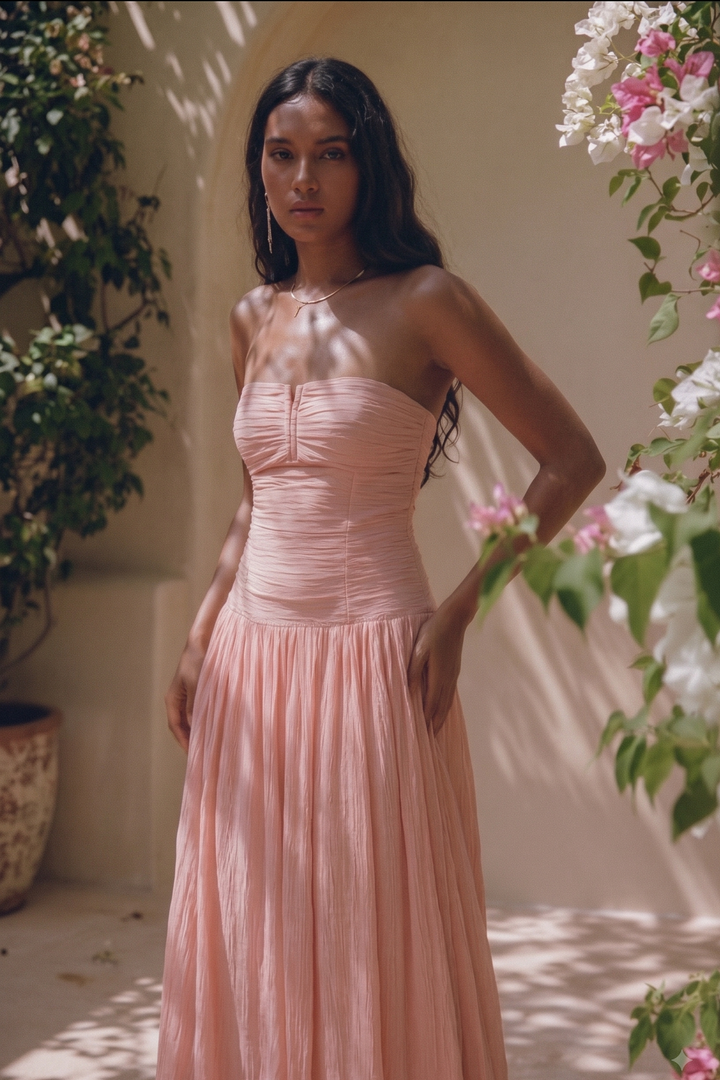 A woman wearing a strapless dusty pink maxi dress with a ruched bodice and flowing skirt, standing in a sunlit outdoor setting with climbing flowers and greenery.