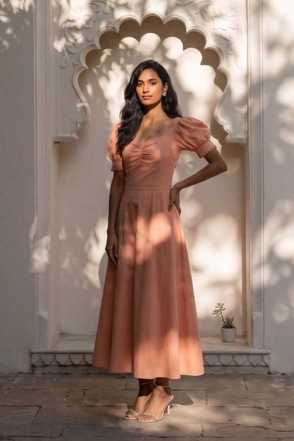A woman stands in front of a white arched architectural wall with leafy shadows cast across her and the scene. She is wearing a dusty rose pink puff-sleeve midi dress with a V-neckline and a flared skirt.