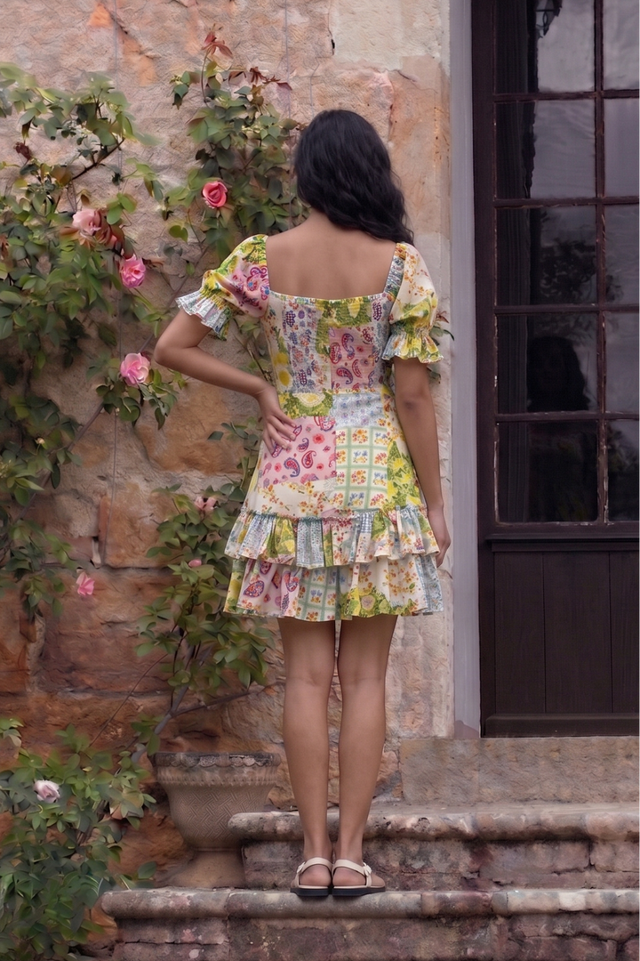 A woman stands on stone steps facing a rustic stone wall with climbing roses and a dark wooden door, wearing a colorful floral tiered mini dress with puff sleeves.