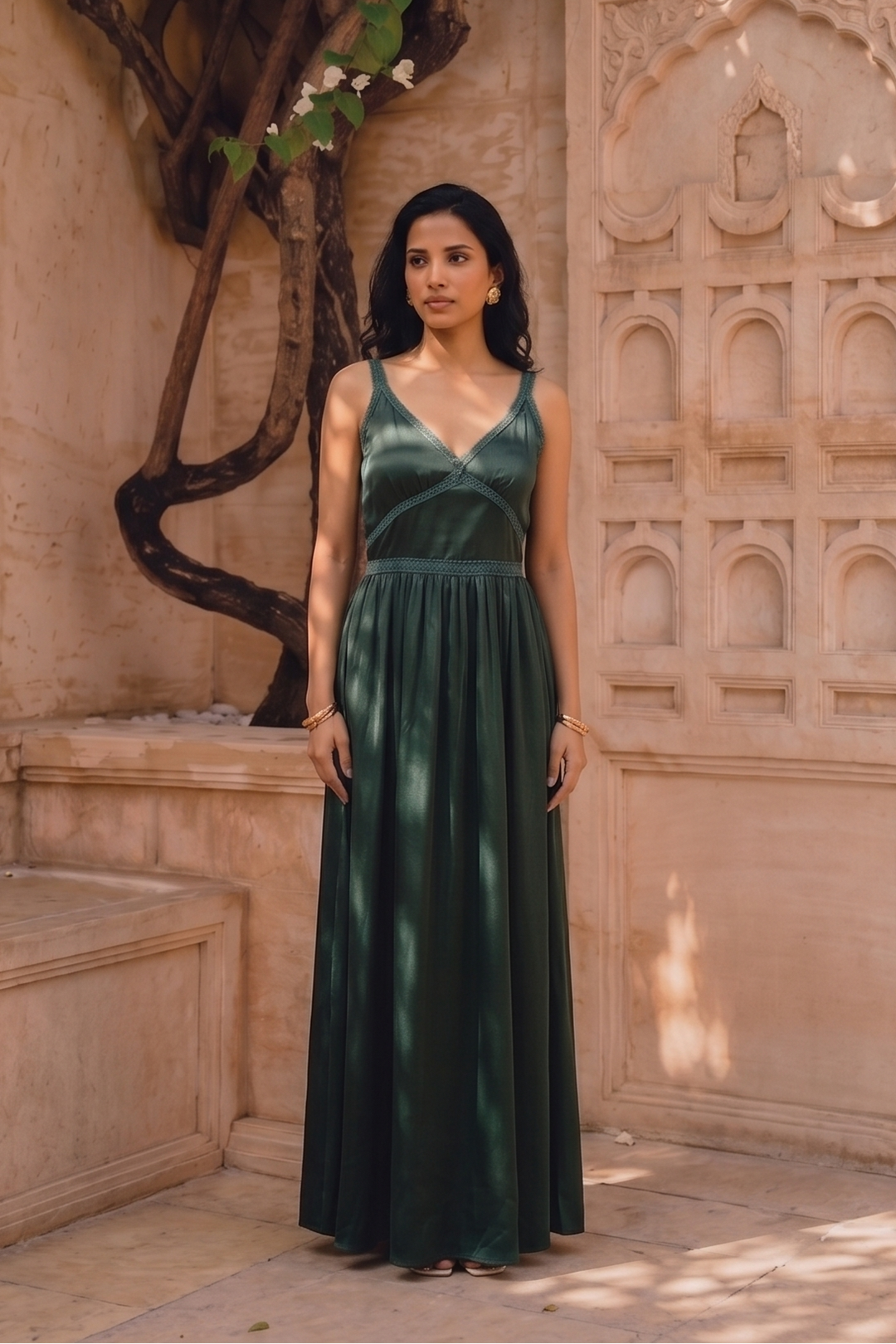 A woman wearing a dark green sleeveless maxi dress stands in a sunlit courtyard with carved beige stone walls and white flowers overhead.