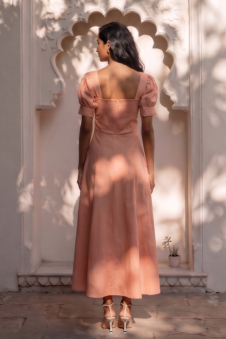A woman stands with her back to the camera in front of an ornate arched wall, wearing a dusty rose pink midi dress with puffed short sleeves and a square neckline.