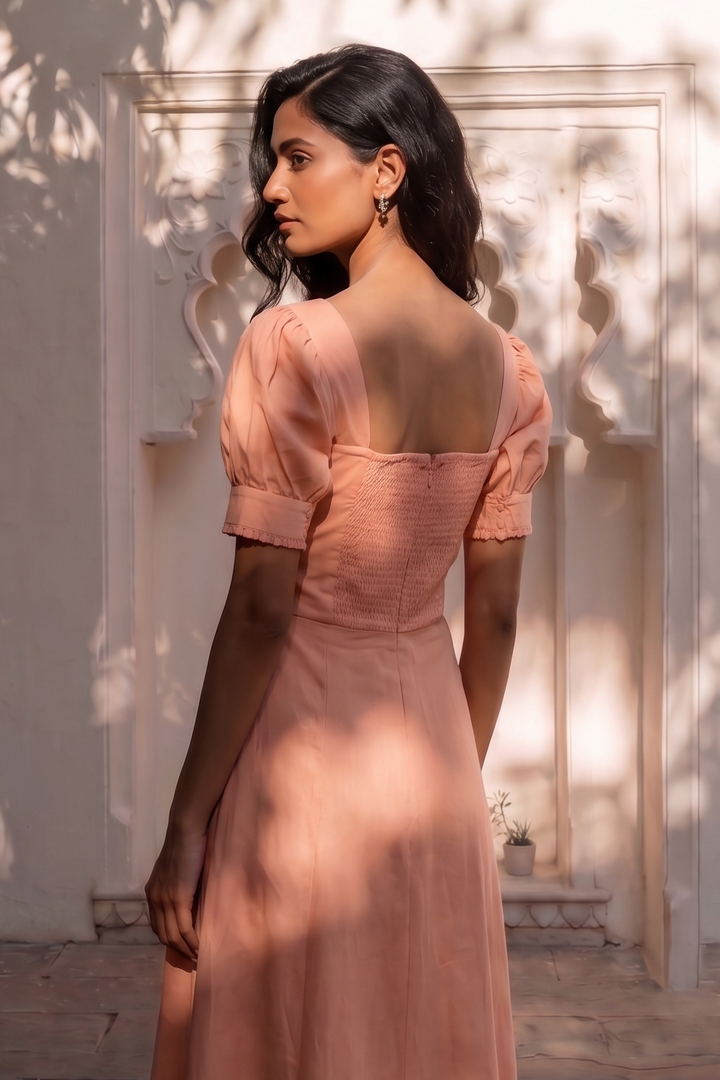 A woman wearing a peach-pink short-sleeve midi dress with a square neckline and backless design, standing in front of a white arched wall with leaf shadows.