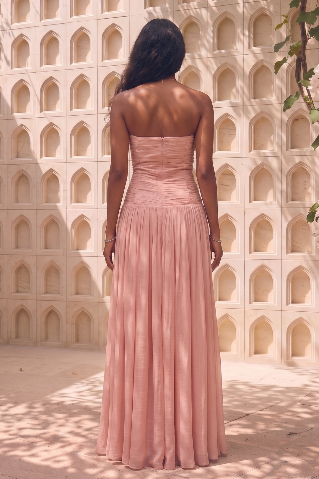 A woman stands with her back to the camera wearing a strapless dusty pink maxi dress with a structured bodice and flowing pleated skirt, set against a sunlit stucco wall with arched recesses and leafy vines.
