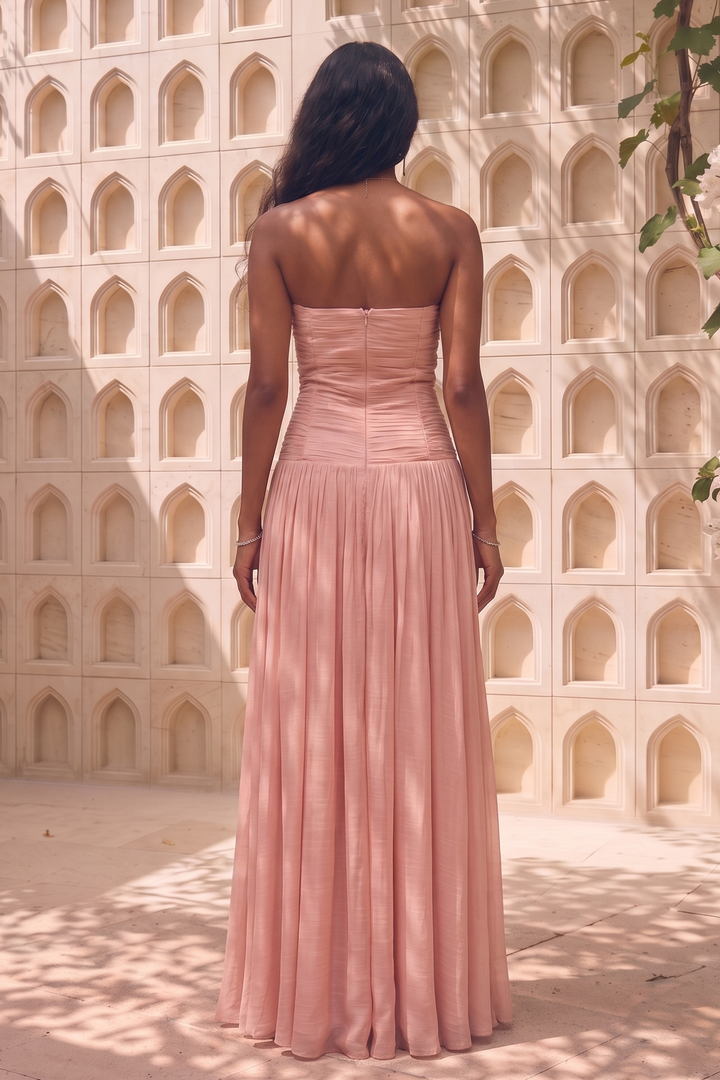 A woman stands with her back to the camera wearing a strapless dusty pink maxi dress with a structured bodice and flowing pleated skirt, set against a sunlit stucco wall with arched recesses and leafy vines.