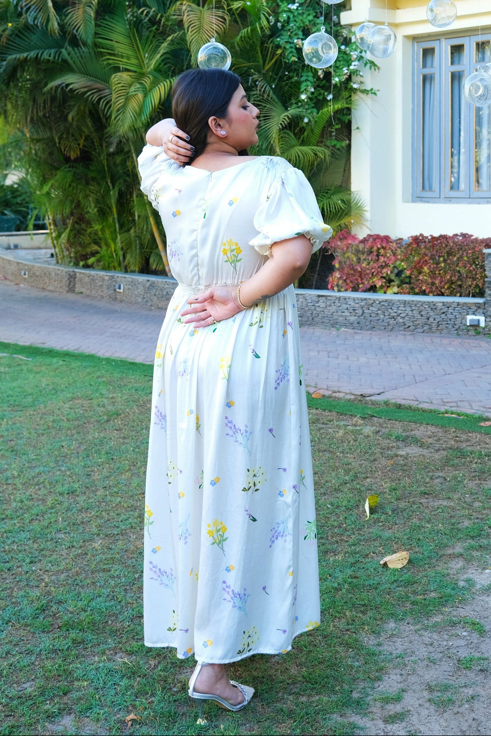 A woman wearing a white puff-sleeve maxi dress with a yellow and purple floral print stands outdoors in a garden setting, looking upward toward floating clear bubbles near a yellow building.