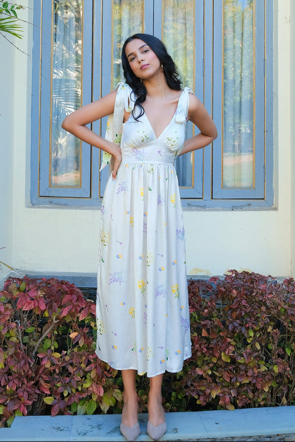 A person wearing a white floral maxi dress with tie-up straps standing in front of a building with large windows.