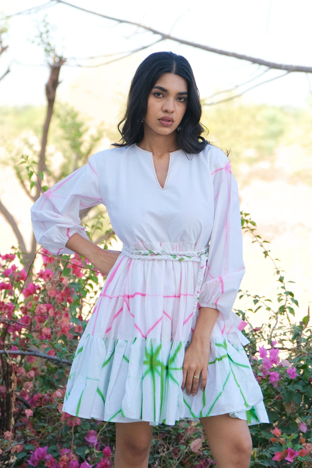 A woman wearing a white mini dress with pink and green abstract line patterns, standing outdoors in front of pink flowers and trees.