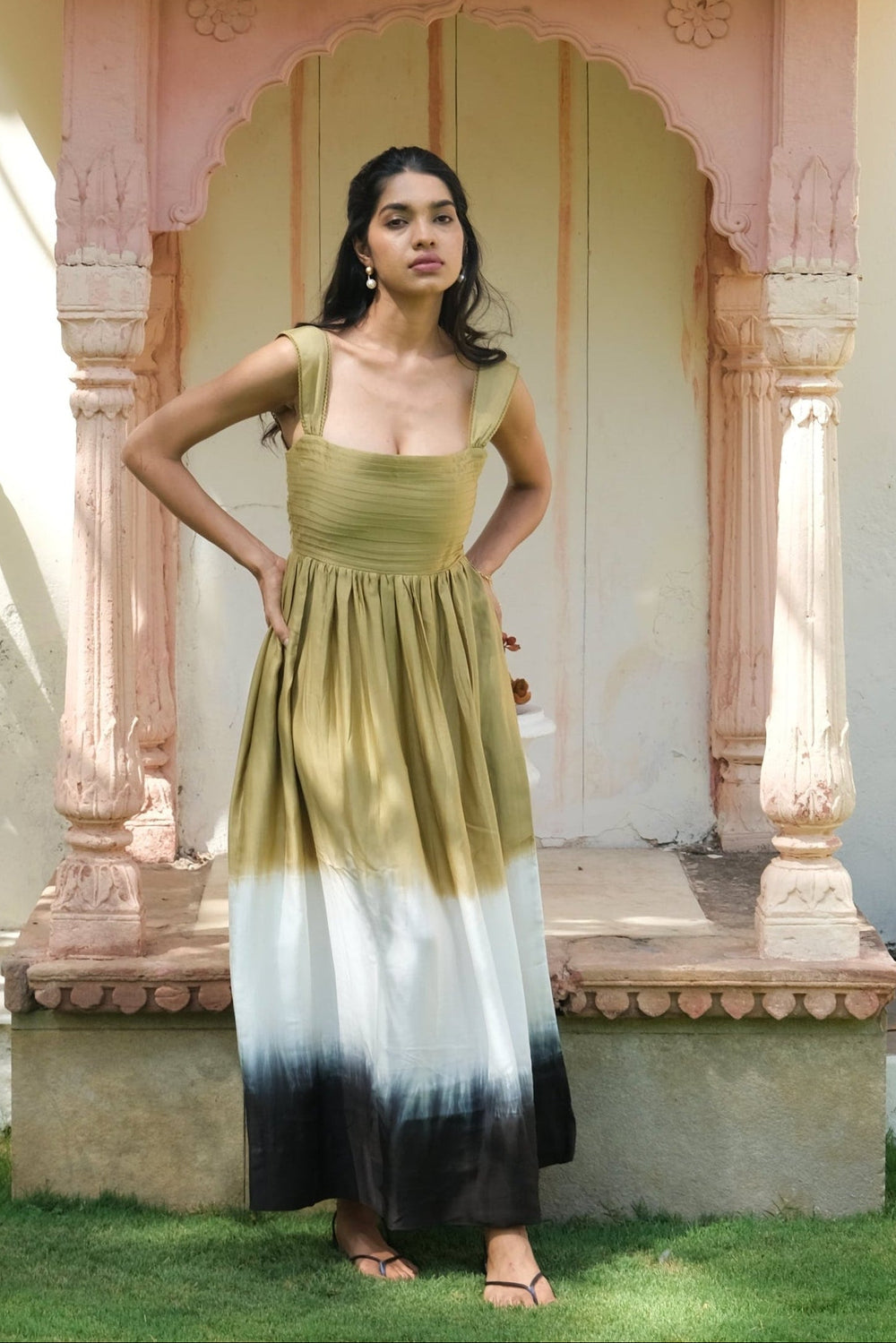 A woman stands outdoors in front of a pink arched doorway, wearing a sleeveless midi dress with a vertical ombré transition from olive green at the top through white to black at the hem.