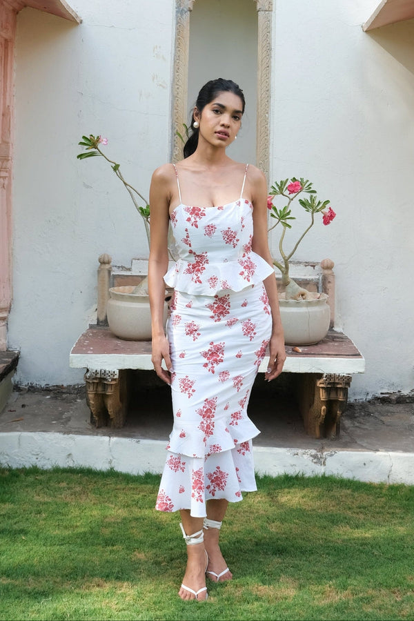 A woman stands outdoors on a grassy lawn in front of a cream-colored building, wearing a white ruffled spaghetti-strap midi dress with a pink floral print. She is wearing white strappy high-heeled sandals.