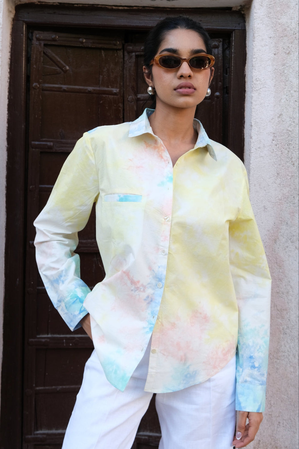 A woman wearing a pastel tie-dye button-down shirt with white pants, standing in front of a rustic beige stucco wall and dark wooden door.