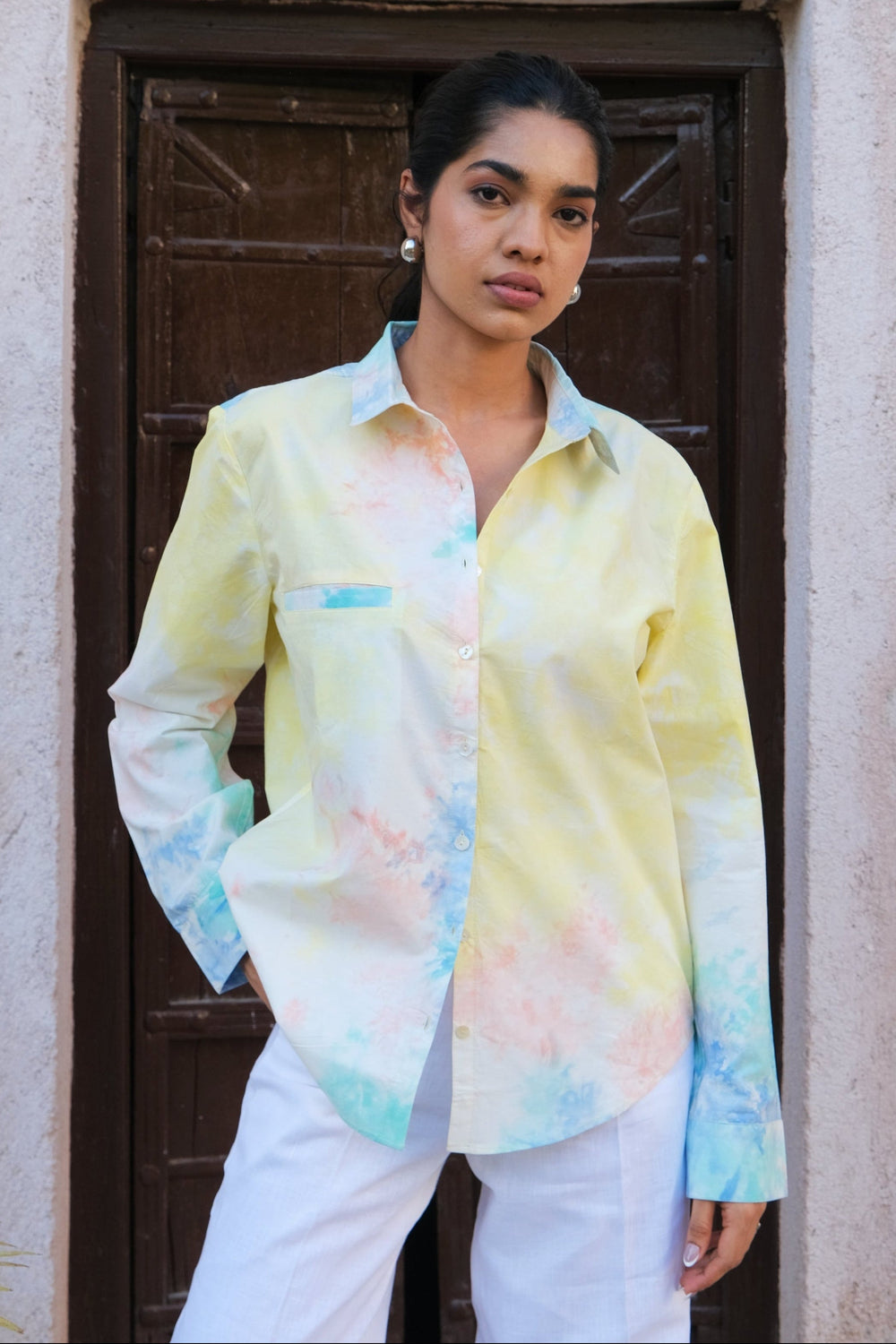 A woman wearing a pastel tie-dye button-up shirt with yellow, blue, and pink tones, paired with white pants, standing in front of a rustic dark brown wooden door and textured beige stucco wall.