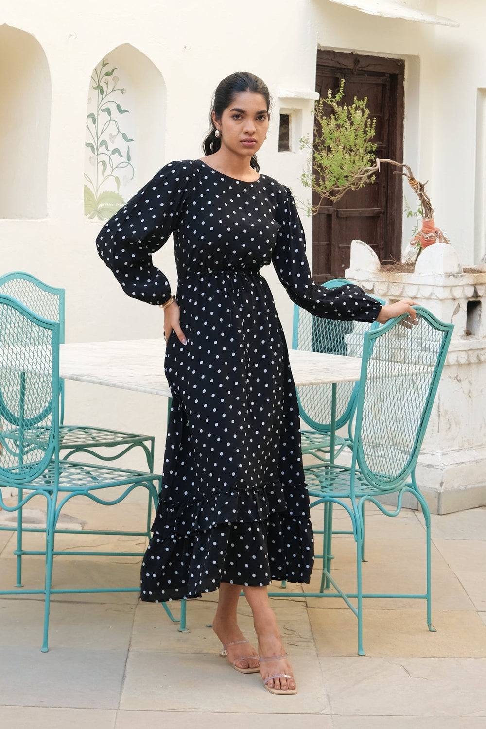 A woman wearing a black long-sleeve maxi dress with white polka dots stands in a courtyard next to turquoise metal chairs and a white table.