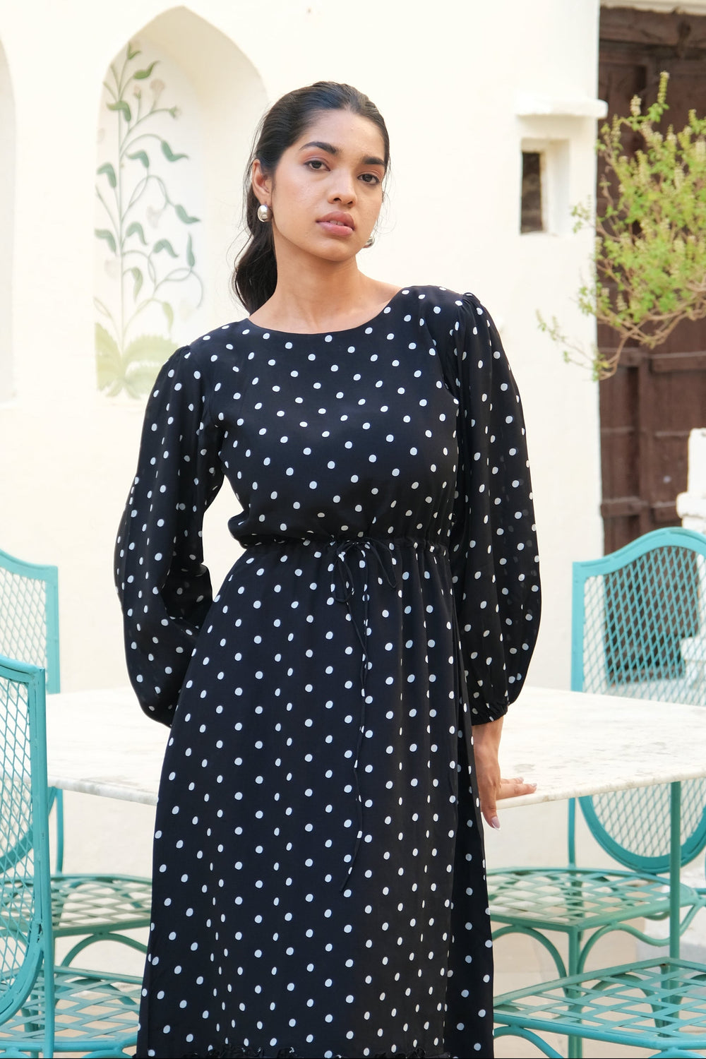 A woman wearing a black long-sleeve maxi dress with white polka dots stands in front of a white stucco wall with arches, a wooden door, and turquoise metal chairs.