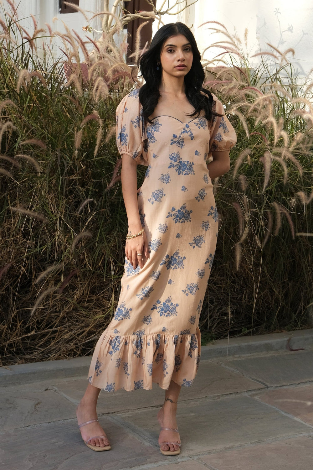 A woman wearing a beige floral maxi dress stands outdoors in front of a white stucco building with arched windows and tall ornamental grasses.