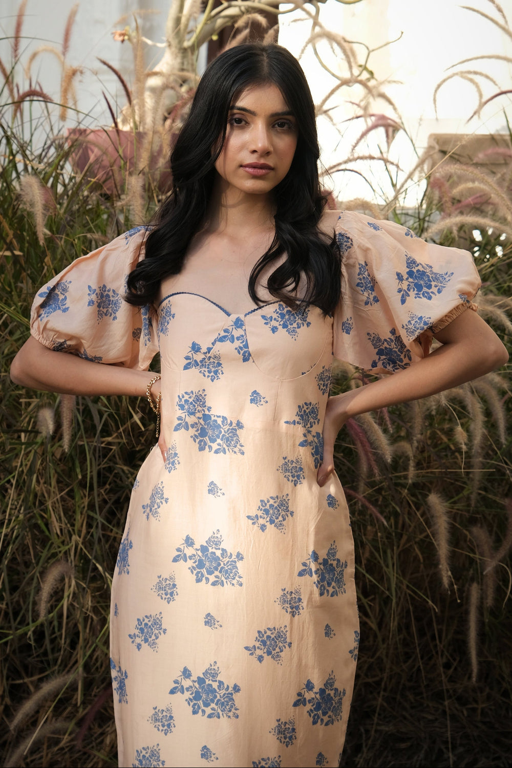 A woman wearing a cream-colored dress with a blue floral pattern and puff sleeves, standing in front of a background of dried ornamental grass.