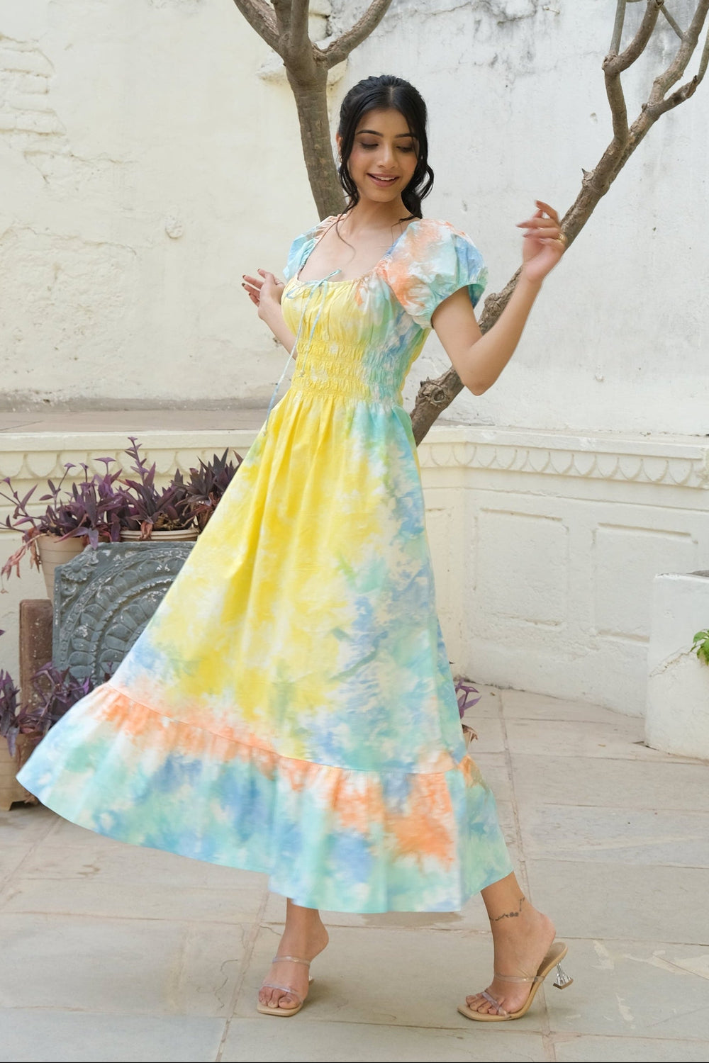 A woman modeling a short-sleeved midi dress with a pastel tie-dye pattern of yellow, blue, and orange, standing outdoors in front of a white stucco wall and leafy tree branches.