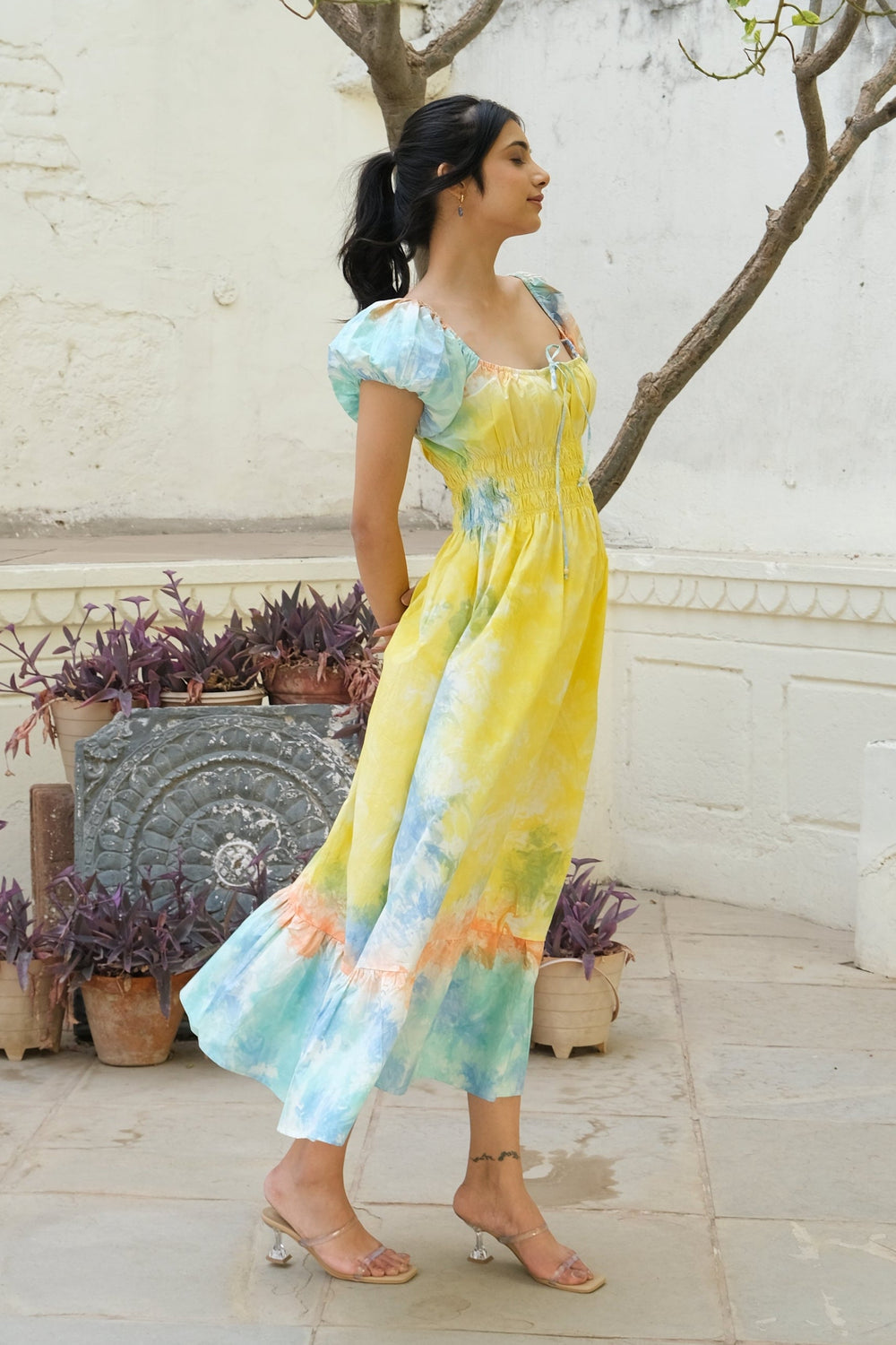 A woman wearing a yellow, light blue, and orange tie-dye midi dress with puff sleeves, standing in an outdoor courtyard with potted plants and a white wall background.