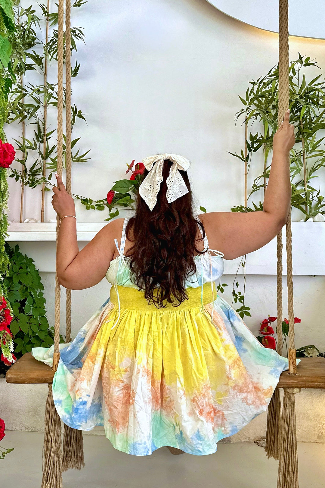 A woman viewed from behind wearing a pastel tie-dye dress with a white bow headband, sitting on a rope swing bench surrounded by greenery and red flowers.