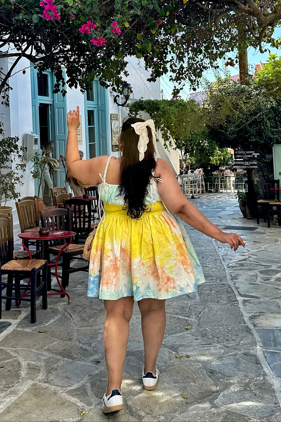 A woman wearing a yellow and blue floral print dress is seen from behind, walking down a stone pathway in a Mediterranean-style outdoor cafe under a canopy of blooming pink bougainvillea flowers.