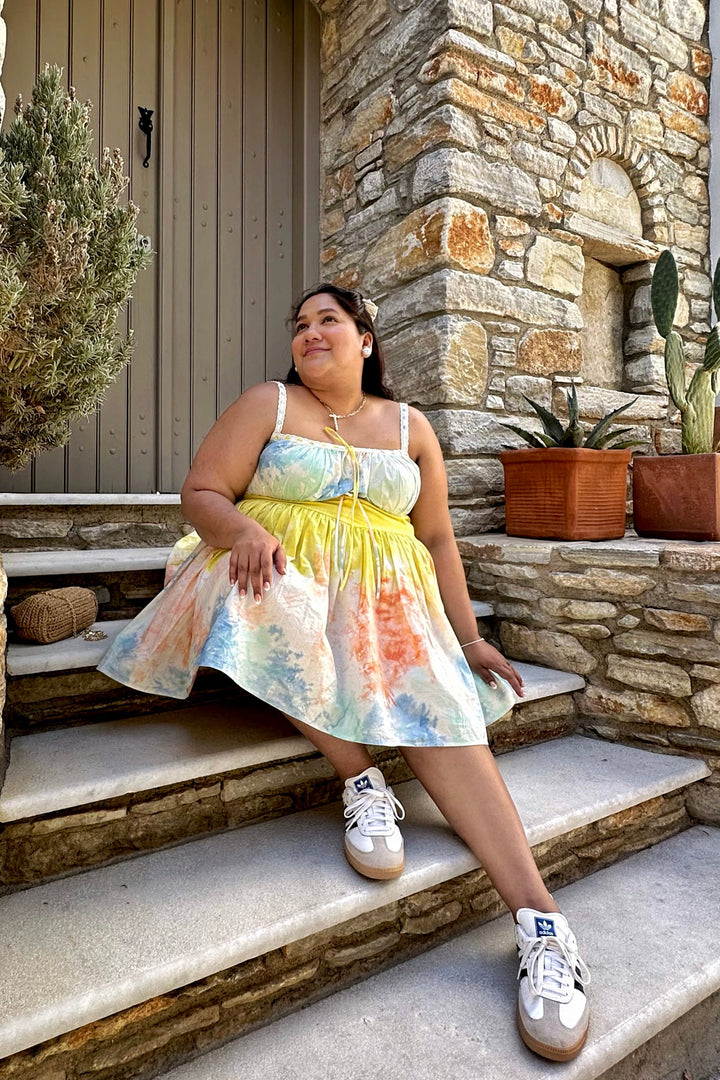 A woman sits on stone steps in front of a rustic stone building, wearing a pastel tie-dye spaghetti strap dress with a yellow bodice and white sneakers.