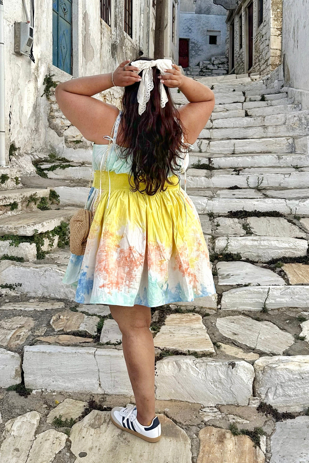 A woman stands on stone stairs in a Mediterranean village, viewed from behind, wearing a tie-dye sleeveless dress with yellow, blue, and orange tones, white sneakers, and a white headscarf tied with a string.