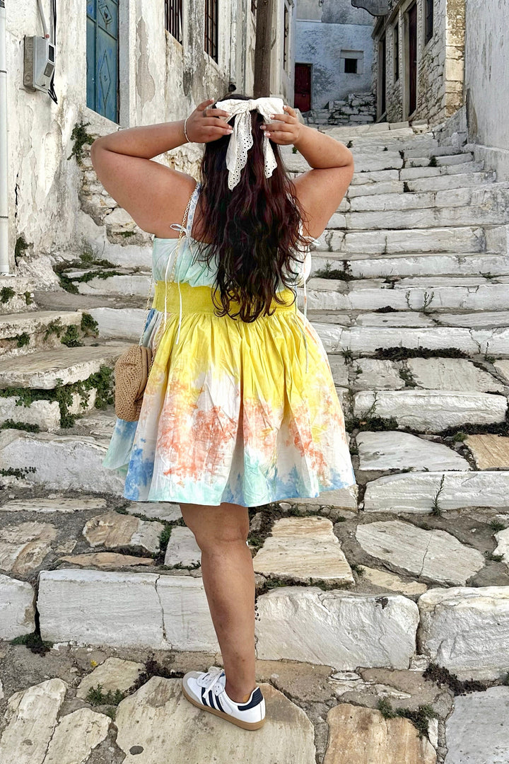 A woman stands on stone stairs in a Mediterranean village, viewed from behind, wearing a tie-dye sleeveless dress with yellow, blue, and orange tones, white sneakers, and a white headscarf tied with a string.