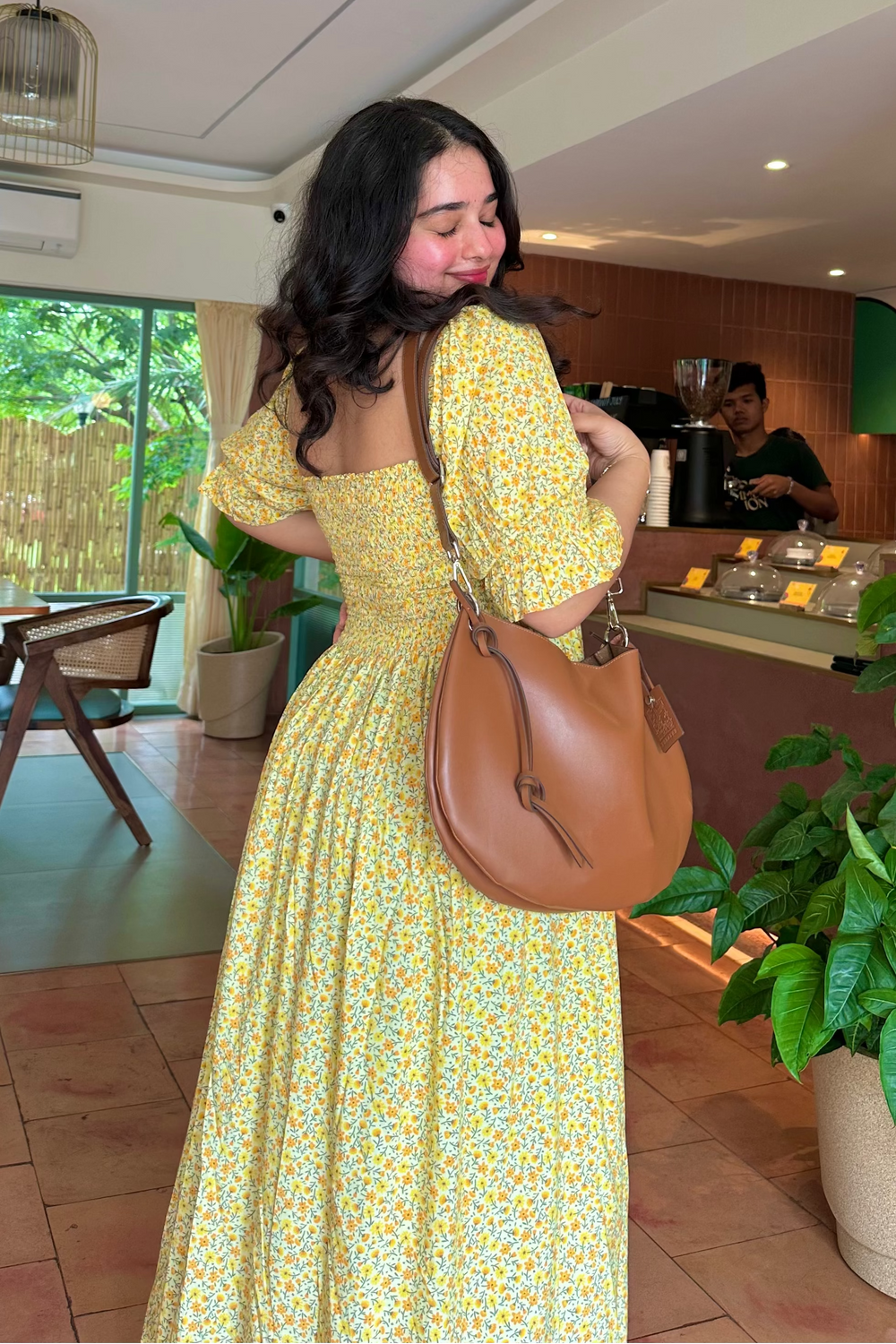 A woman wearing a yellow floral print maxi dress and carrying a brown leather shoulder bag, standing inside a cafe with wooden tables, potted plants, and a counter in the background.