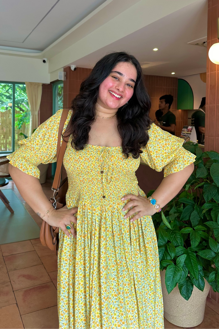 A woman wearing a yellow and white floral print dress with puff sleeves and a tie-neck detail, standing in a cafe-like environment with a potted plant and tiled floor.