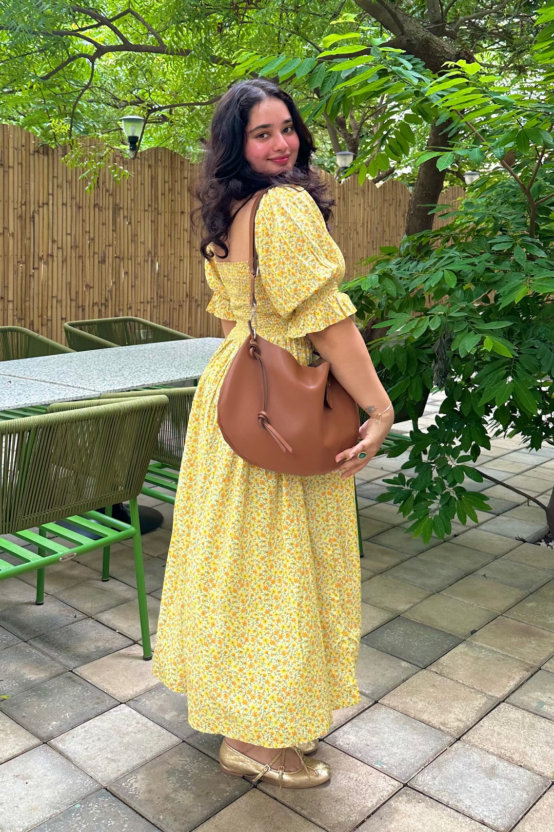 A woman wearing a yellow floral puff-sleeve maxi dress and carrying a brown leather shoulder bag, standing on a tiled patio in front of a bamboo fence and green trees.
