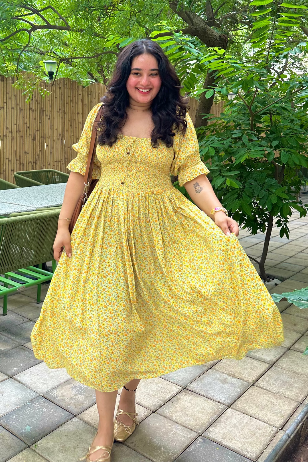 A woman wearing a yellow floral midi dress stands outdoors on a paved patio area surrounded by green trees and garden furniture.