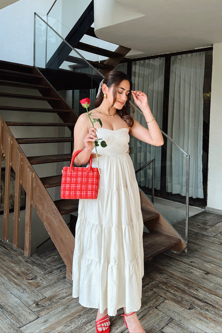 A woman wearing a white tiered maxi dress stands in front of a staircase holding a red rose and carrying a red plaid handbag.