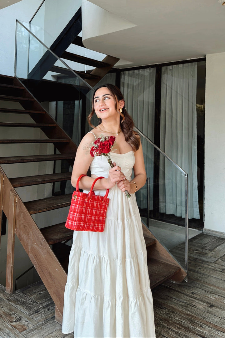 A woman wearing a white strapless tiered maxi dress stands in front of a modern wooden staircase, holding a red plaid handbag and a bouquet of red roses.