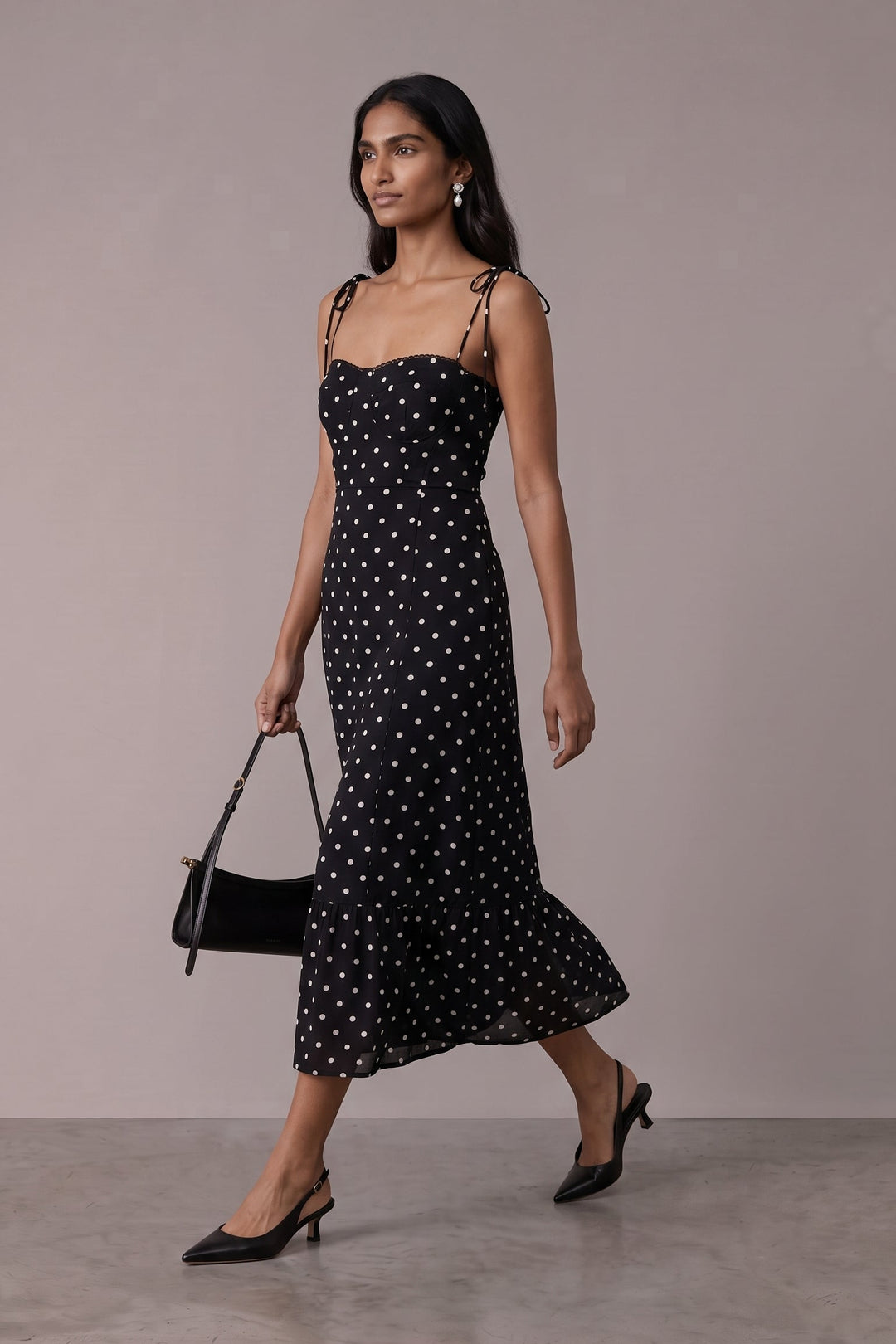 A woman models a black midi dress with white polka dots and thin shoulder straps, walking against a neutral studio backdrop while holding a black handbag.