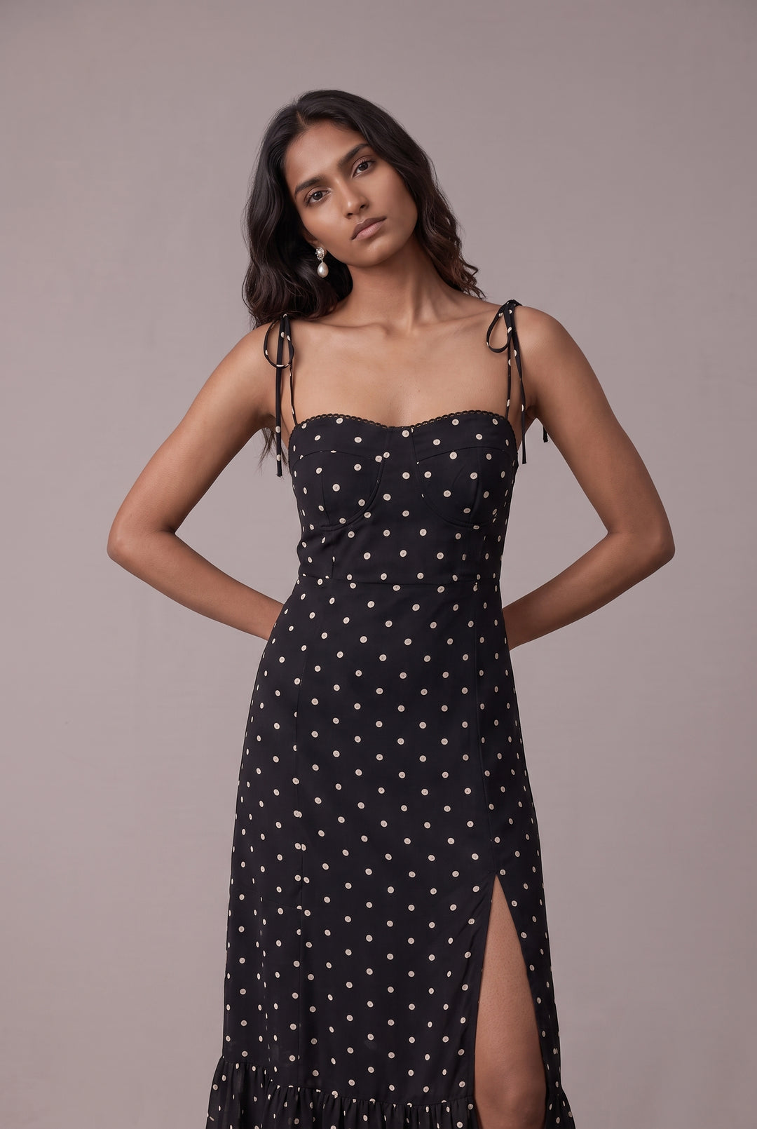 A woman modeling a black midi dress with white polka dots, featuring thin shoulder ties and a thigh-high side slit, set against a solid taupe background.