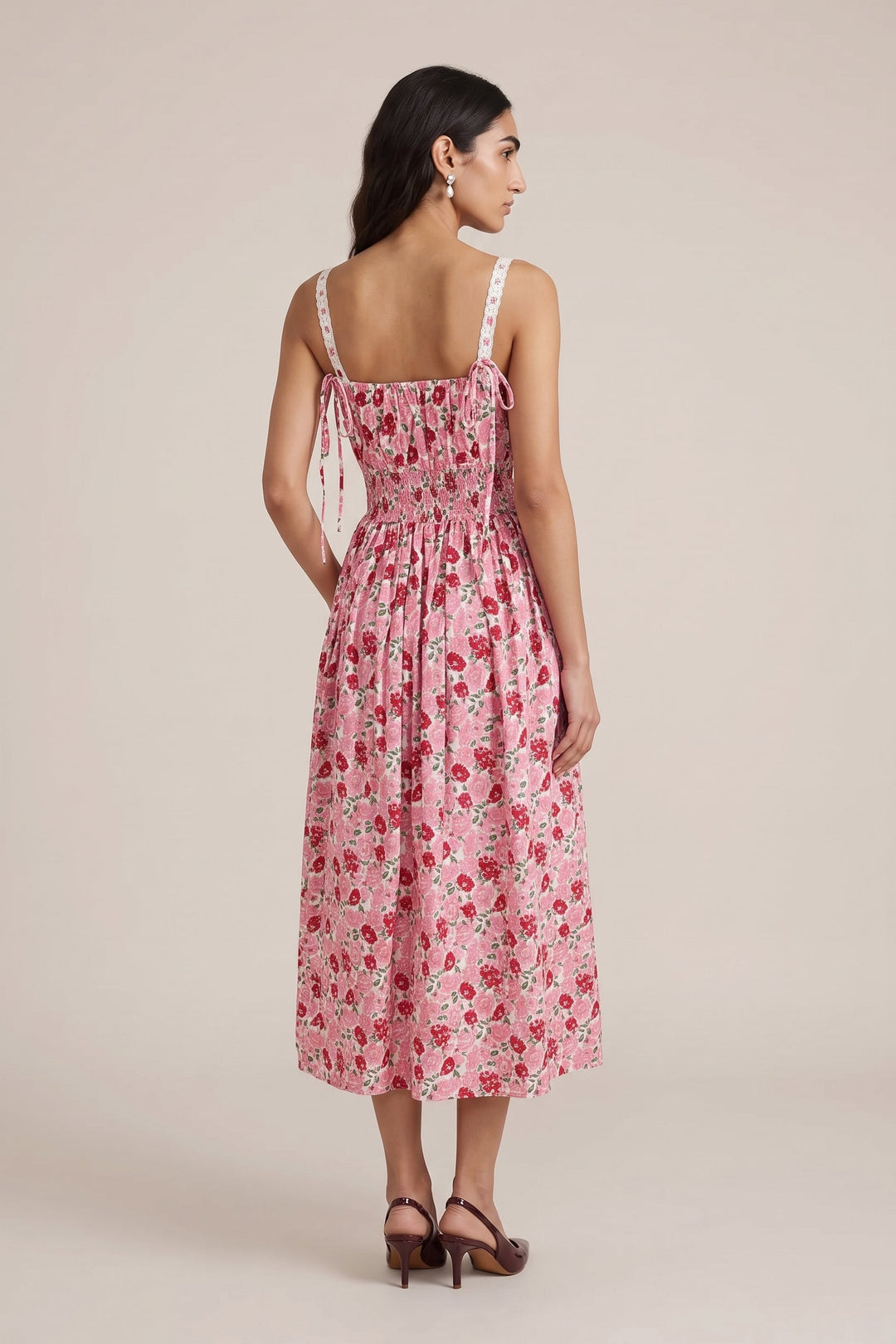 A woman modeling a pink floral midi dress with white tie-shoulder straps, shown from the back against a solid beige studio background.