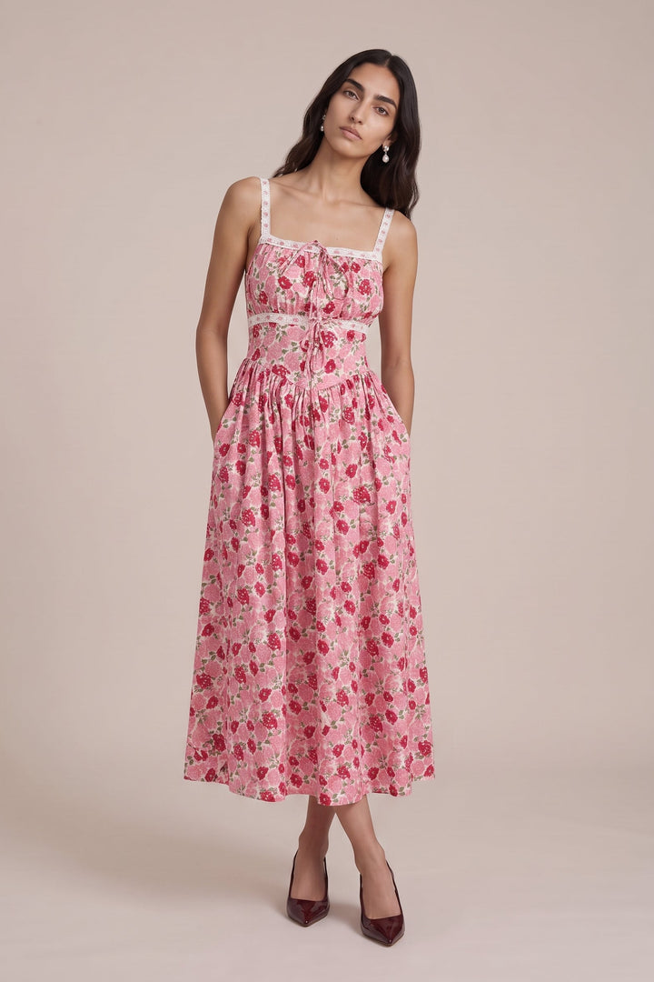 A woman modeling a pink midi dress with a red floral print, featuring thin straps, a square neckline with a bow detail, and side pockets, set against a solid beige studio background.