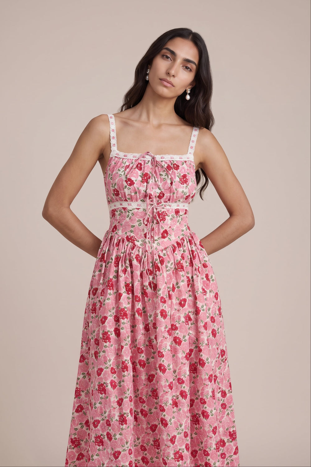 A woman modeling a pink floral midi dress with a tie-neck detail and thin straps, set against a solid beige studio background.