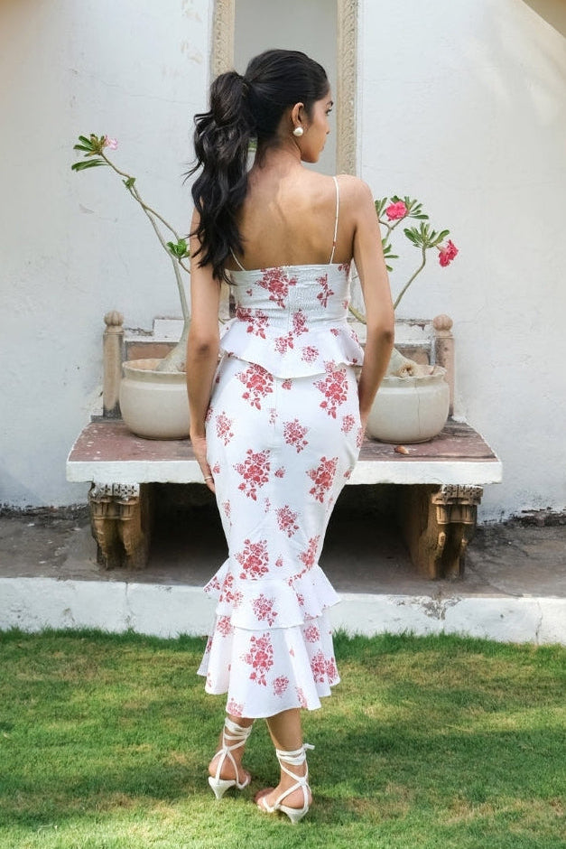 A woman stands outdoors in front of a cream-colored building, wearing a white spaghetti-strap maxi dress with a pink floral print and a ruffled hem. She is viewed from behind, standing on a patch of green grass.