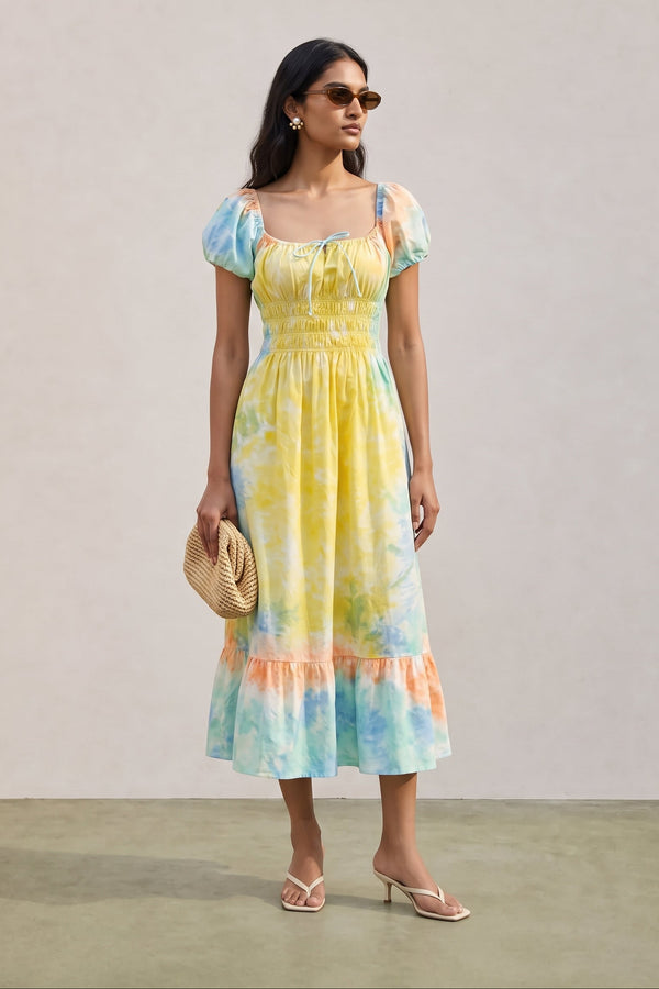 A woman modeling a pastel tie-dye midi dress with puff sleeves, a smocked bodice, and a ruffled hem. The dress features a blend of yellow, blue, and peach tones.