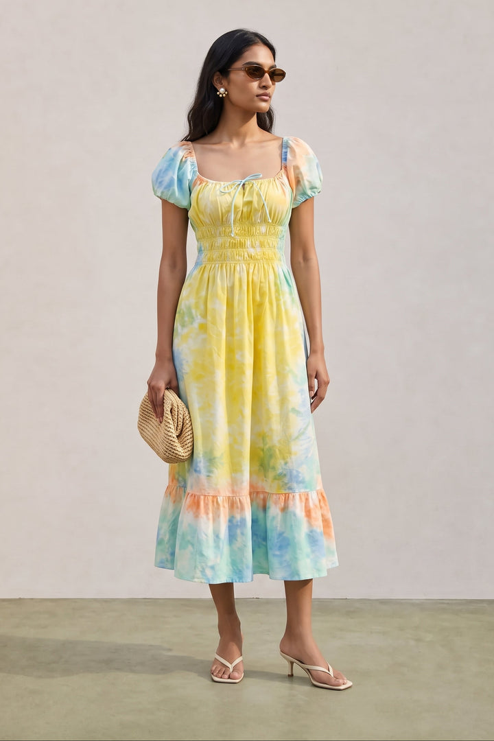A woman modeling a pastel tie-dye midi dress with puff sleeves, a smocked bodice, and a ruffled hem. The dress features a blend of yellow, blue, and peach tones.